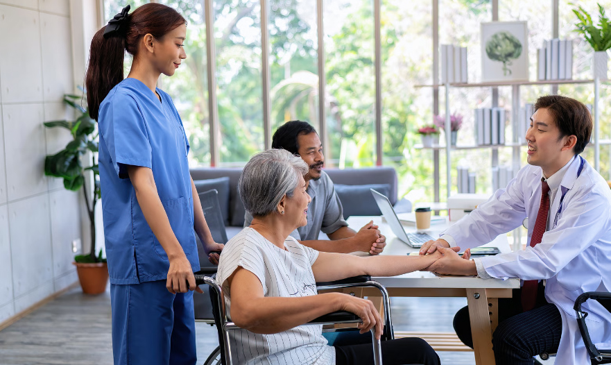 A smiling male doctor in a white coat sits at a desk and holds the hand of an elderly woman in a wheelchair during a consultation. A female nurse in blue scrubs stands behind the wheelchair, and another man sits at the table, all in a bright, modern office with large windows.